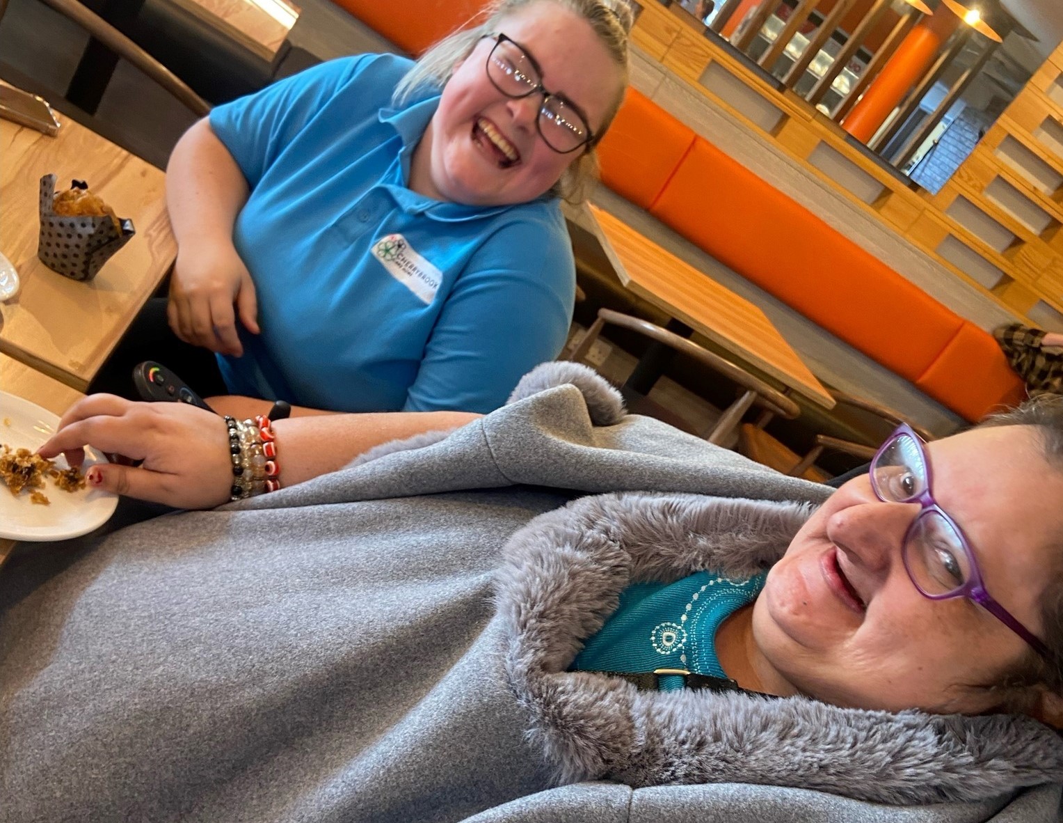 Two women sitting at a wooden table in a café, smiling and laughing. One is wearing a light blue uniform with a care home logo and glasses, while the other is draped in a grey coat with a fur-trimmed collar, wearing purple glasses and a teal top. A plate with crumbs and a muffin in a metal holder is visible on the table, and the orange seating and wooden décor of the café are in the background