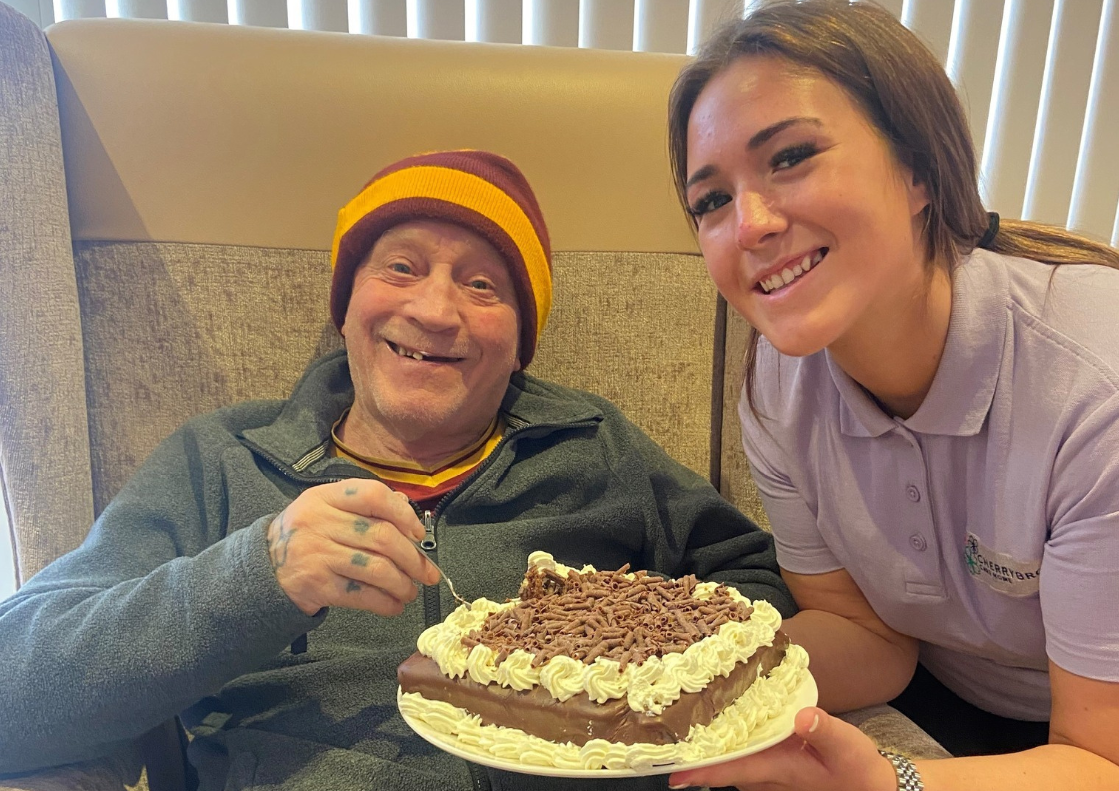 A happy resident at Cherrybrook Care Home, wearing a maroon and gold beanie, enjoying a slice of chocolate cake with cream, alongside a smiling caregiver in a light purple uniform.